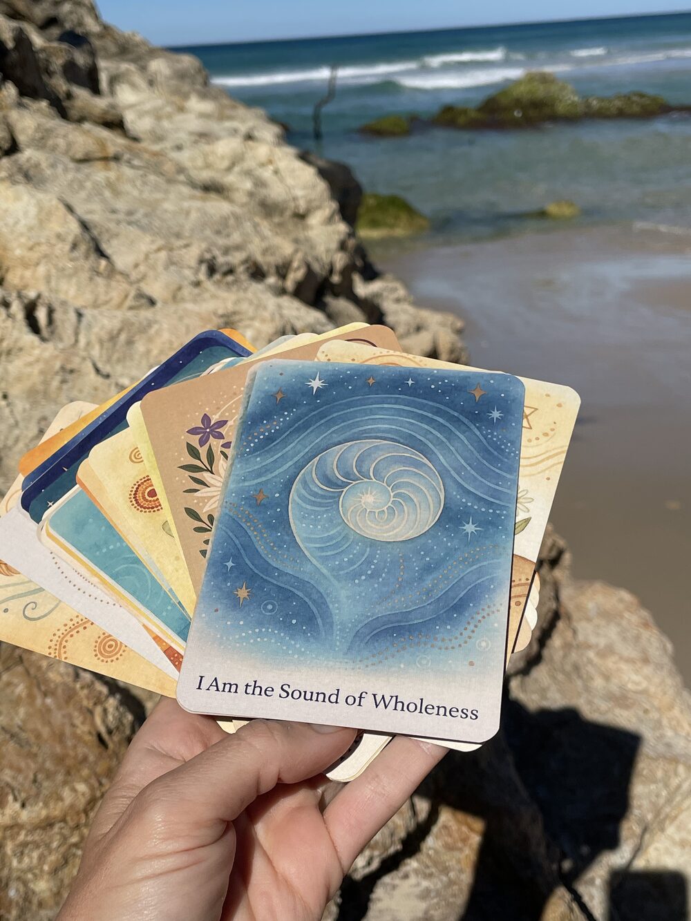 Hand holding a fan of Rainbow Earth cards on ocean rocks, top card reading I Am the Sound of Wholeness